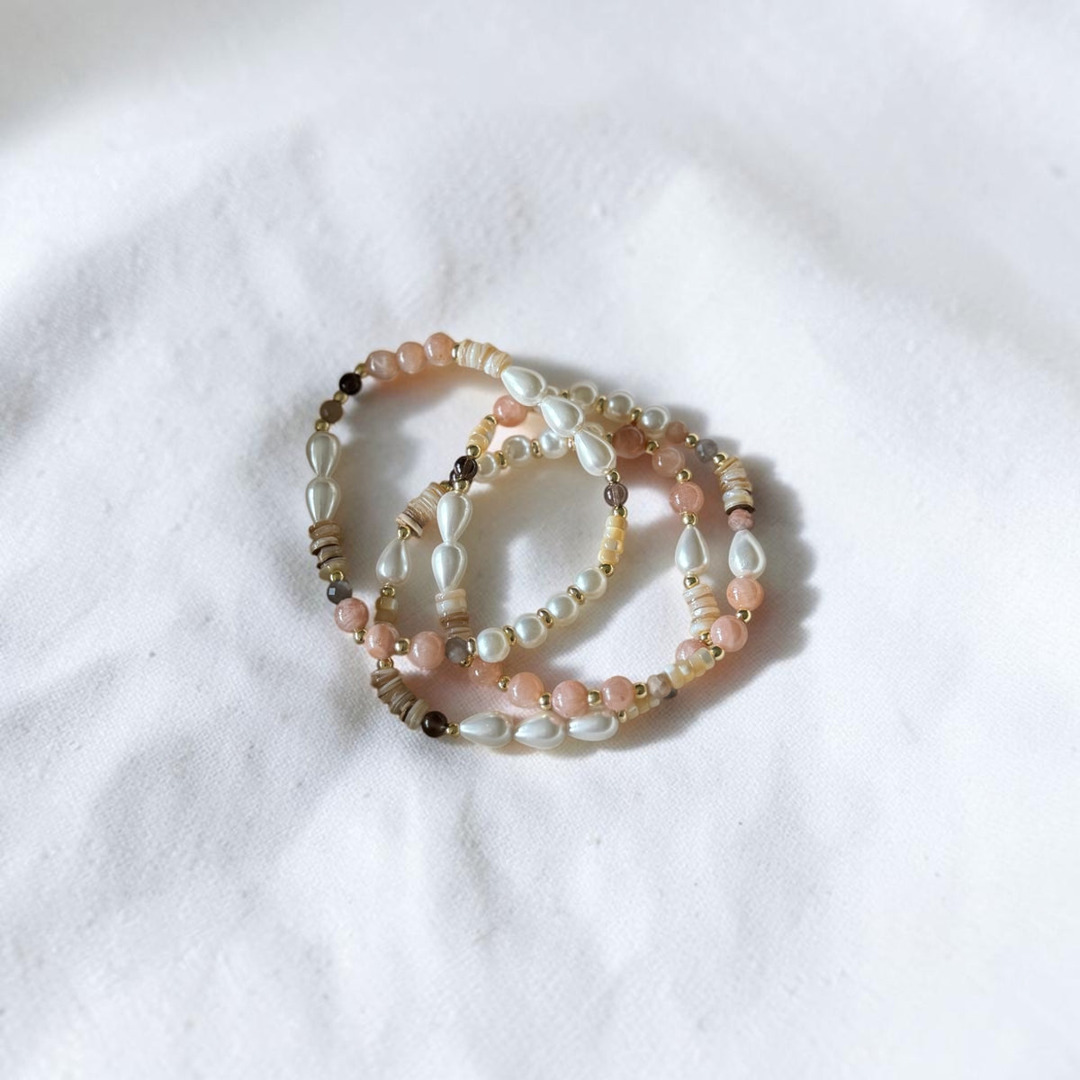 Three beaded stretch bracelets with pink, white, and metallic gold beads, arranged in a circle on a textured white fabric background.