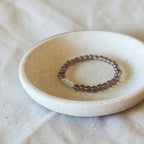 A beaded stretch ring with small brown beads and a clear faceted accent stone, displayed in a small round ceramic dish on a textured beige fabric background.