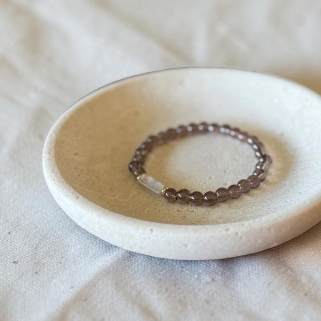 A beaded stretch ring with small brown beads and a clear faceted accent stone, displayed in a small round ceramic dish on a textured beige fabric background.