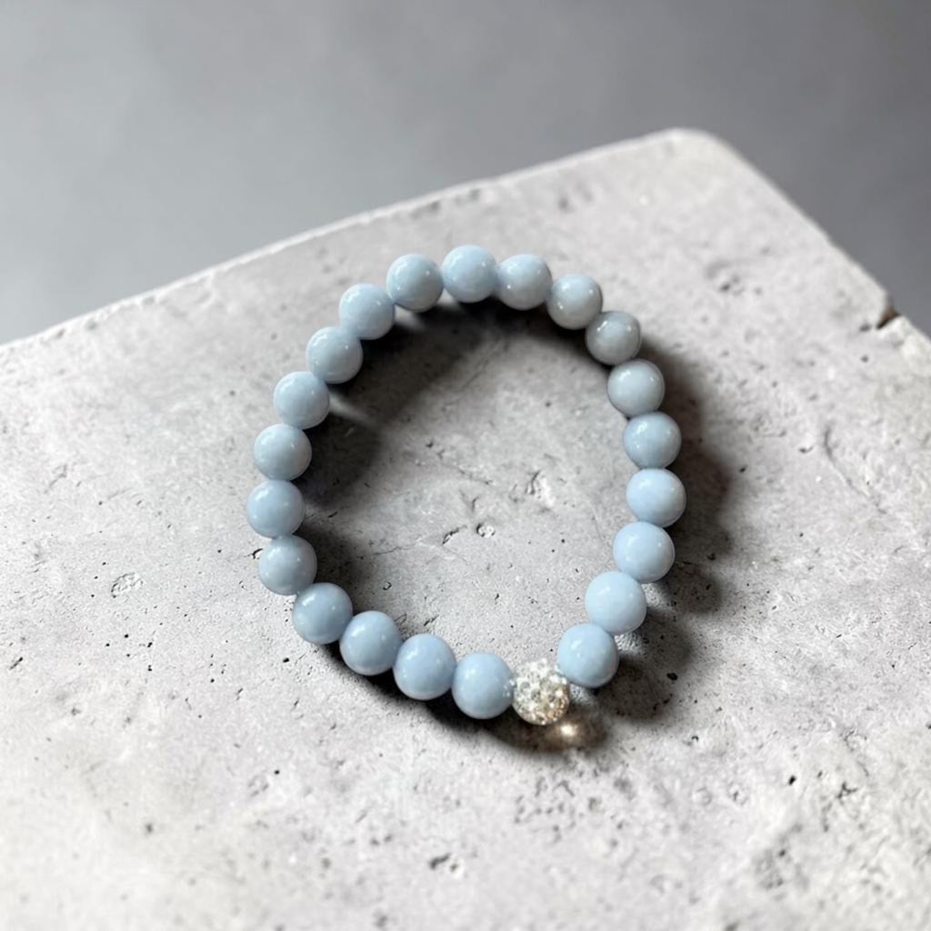 A light blue beaded bracelet with a small silver-toned rhinestone accent bead, resting on a textured gray concrete surface.