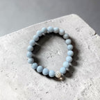A light blue beaded bracelet with a small silver-toned rhinestone accent bead, resting on a textured gray concrete surface.
