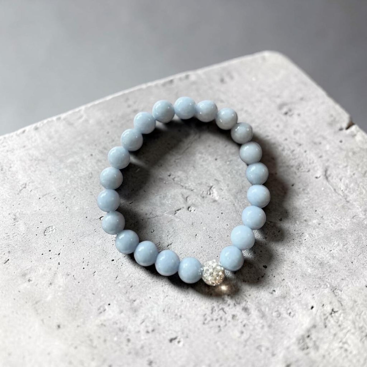 A light blue beaded bracelet with a small silver-toned rhinestone accent bead, resting on a textured gray concrete surface.