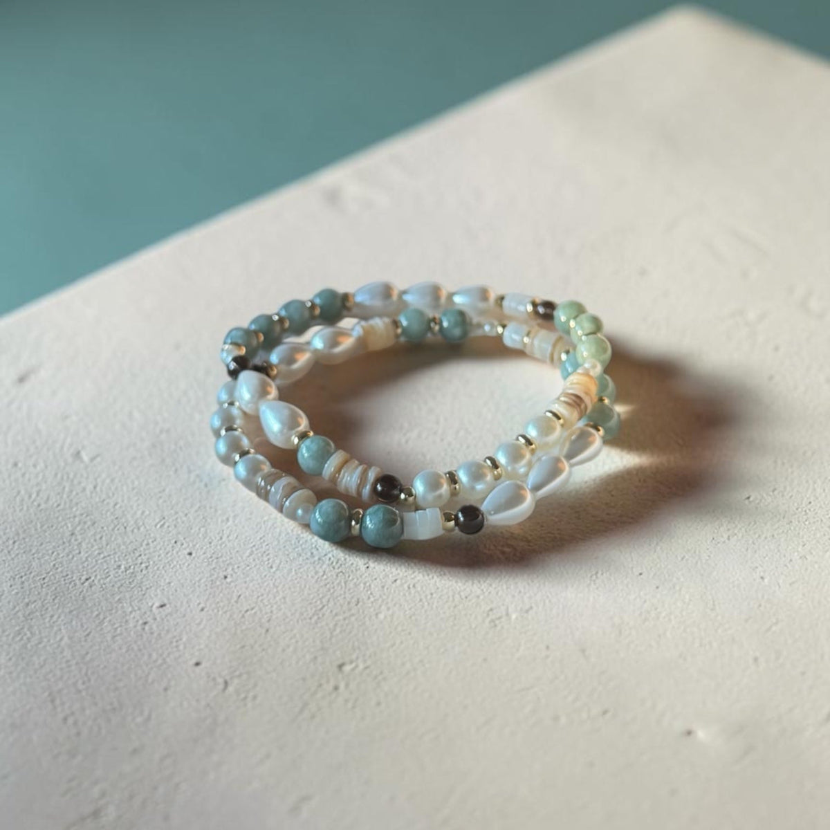 A pair of beaded stretch bracelets with light blue, white, and gray beads, resting on a textured off-white surface with a soft teal background.