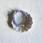 A beaded bracelet made of light purple lavender beads with a single faceted gold accent bead, displayed on a white scalloped ceramic dish against a textured white background.
