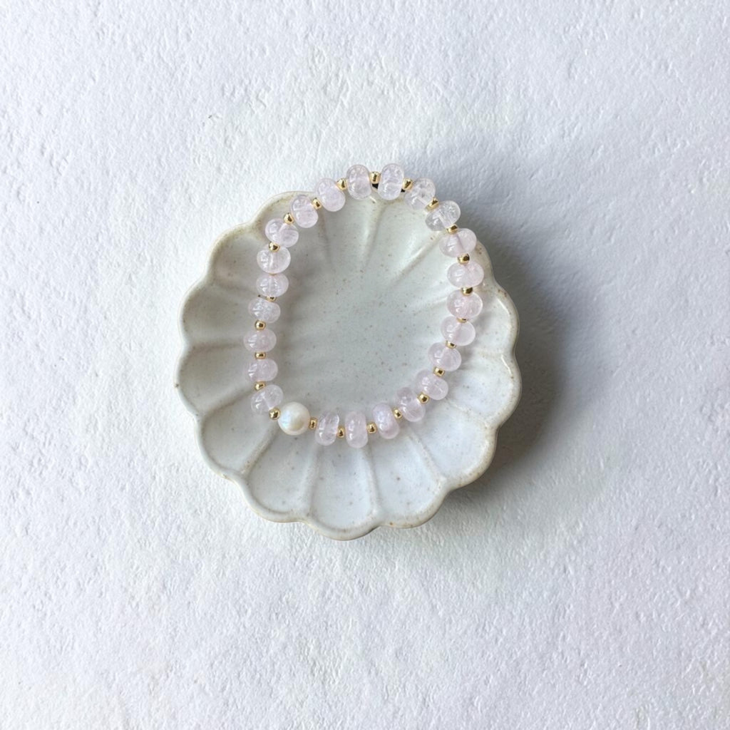 A beaded stretch bracelet made of pale pink translucent beads and small gold spacer beads, displayed on a scalloped off-white ceramic dish against a textured white background.