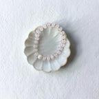 A beaded stretch bracelet made of pale pink translucent beads and small gold spacer beads, displayed on a scalloped off-white ceramic dish against a textured white background.
