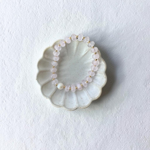 A beaded stretch bracelet made of pale pink translucent beads and small gold spacer beads, displayed on a scalloped off-white ceramic dish against a textured white background.