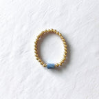 A gold beaded stretch ring bracelet with a single textured blue cylindrical bead, displayed on a textured white background.