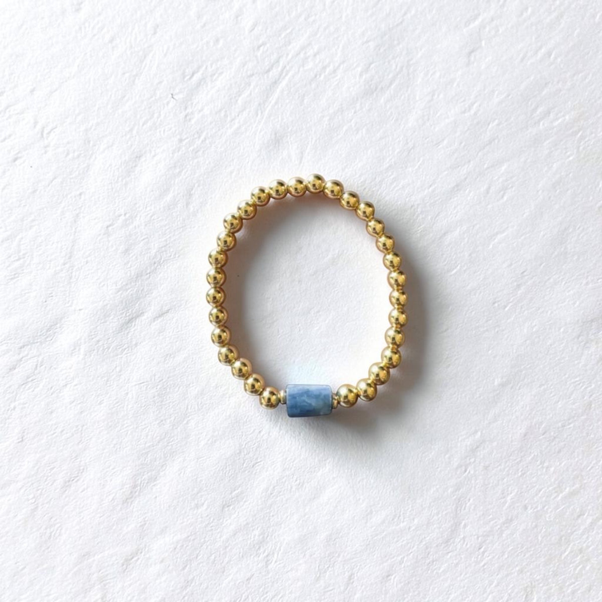 A gold beaded stretch ring bracelet with a single textured blue cylindrical bead, displayed on a textured white background.