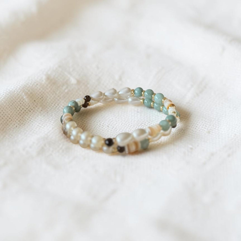 A beaded stretch bracelet bracelet featuring light green, white, and beige beads, resting on a textured white fabric background.