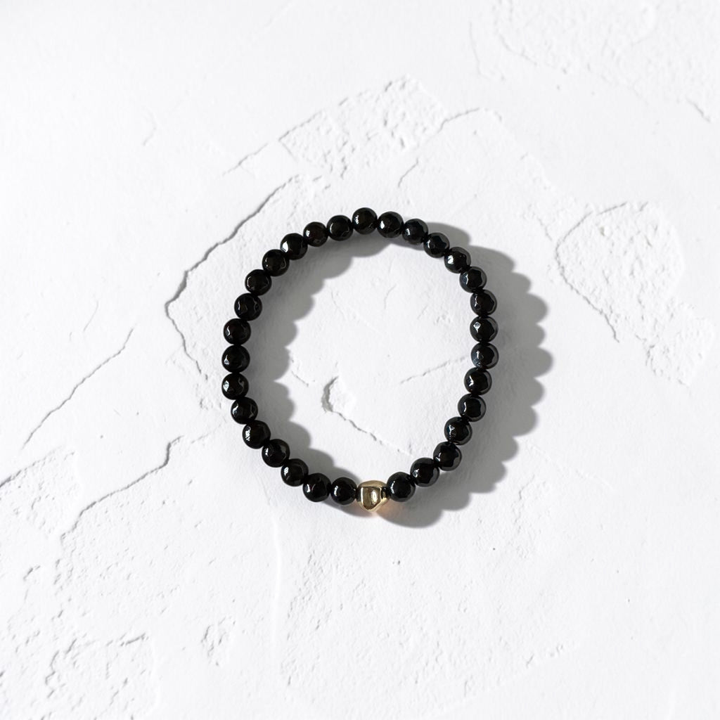 A black beaded stretch bracelet with a small gold accent bead, displayed on a textured white surface.