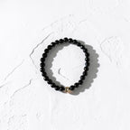 A black beaded stretch bracelet with a small gold accent bead, displayed on a textured white surface.