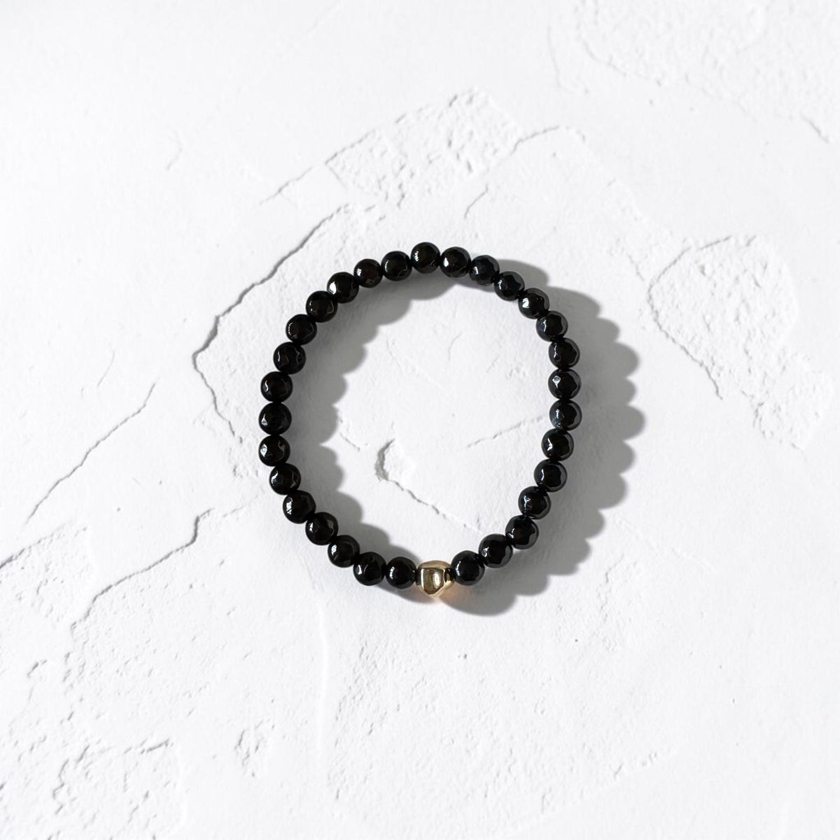 A black beaded stretch bracelet with a small gold accent bead, displayed on a textured white surface.