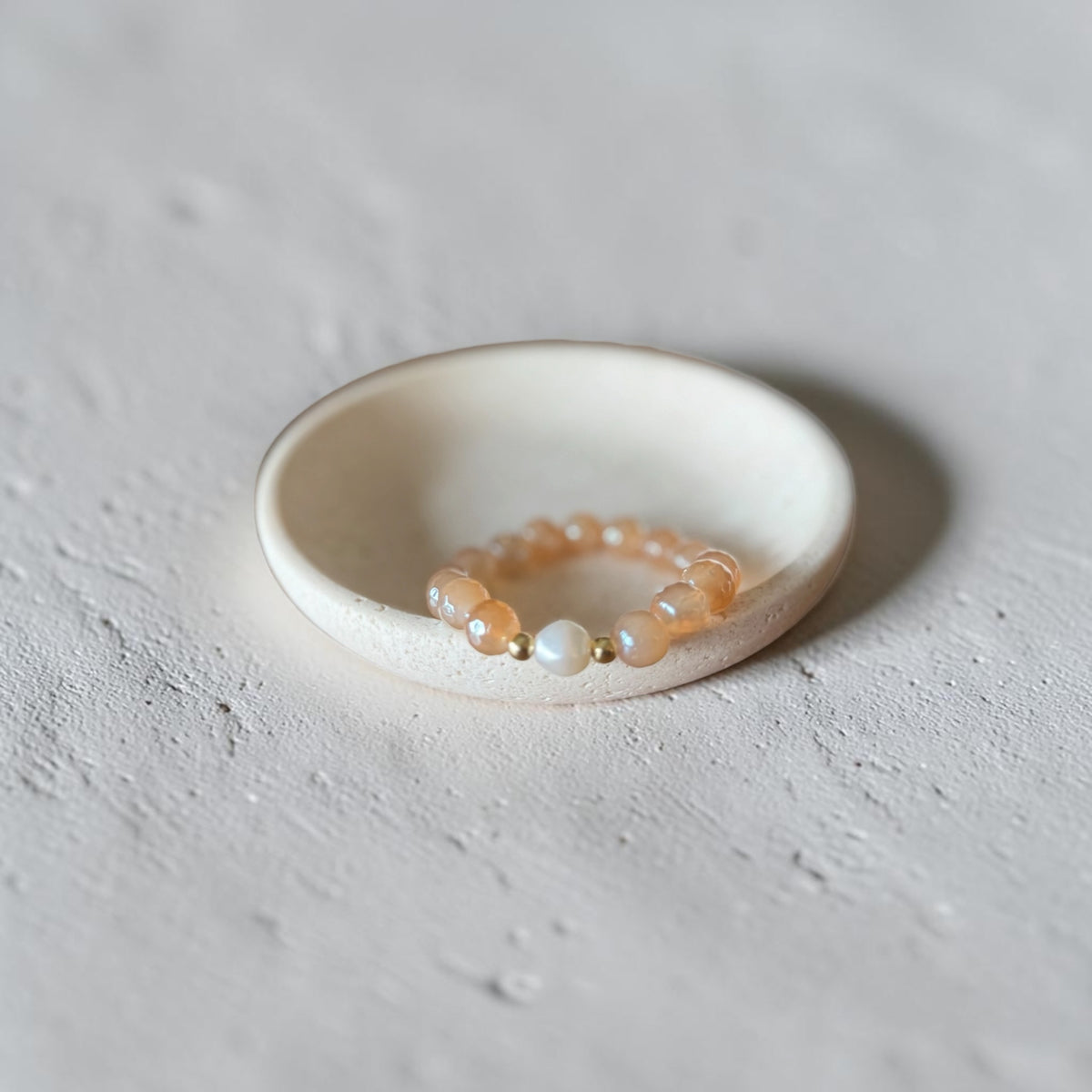 A delicate beaded stretch bracelet with peach-orange translucent beads, a white pearl, and small gold spacer beads, displayed on a small white ceramic dish against a textured light gray background.