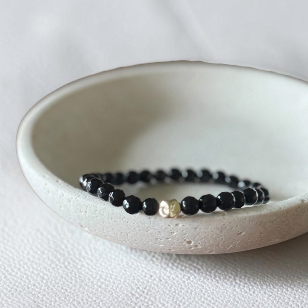 A beaded stretch bracelet with glossy black faceted beads and a small gold-toned accent bead, displayed inside a shallow off-white ceramic dish on a light textured fabric surface.