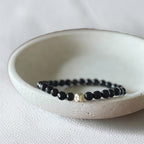 A beaded stretch bracelet with glossy black faceted beads and a small gold-toned accent bead, displayed inside a shallow off-white ceramic dish on a light textured fabric surface.