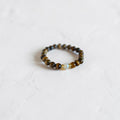 A beaded stretch bracelet featuring brown and black tiger's eye beads with a small light blue stone accent, resting on a textured white surface.