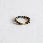 A beaded stretch bracelet featuring brown and black tiger's eye beads with a small light blue stone accent, resting on a textured white surface.