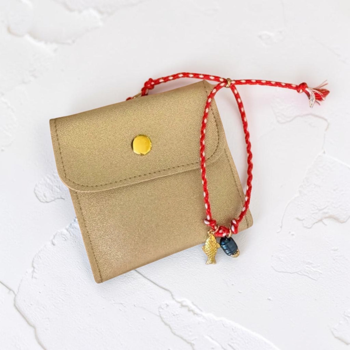 A small tan leather pouch with a gold snap button, featuring a red braided string charm with gold and dark blue beads, photographed on a white textured background.