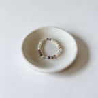 A beaded stretch bracelet with small brown, cream, and white beads, displayed inside a small speckled ceramic dish on a light gray background.