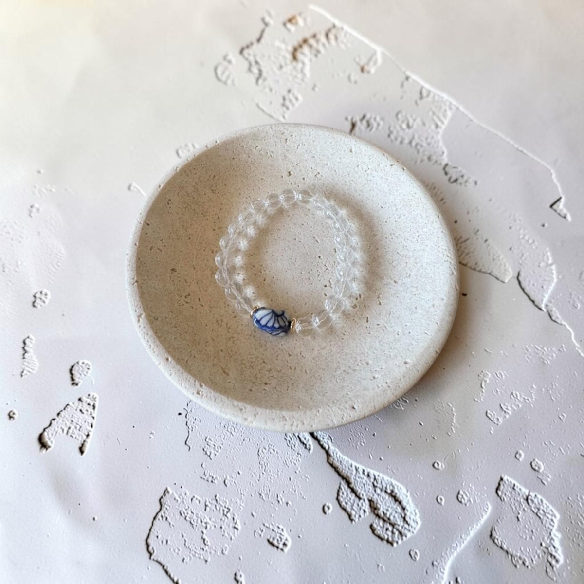 A small beaded bracelet with clear round beads and a blue and white patterned accent bead, displayed inside a round speckled ceramic dish on a textured off-white surface.