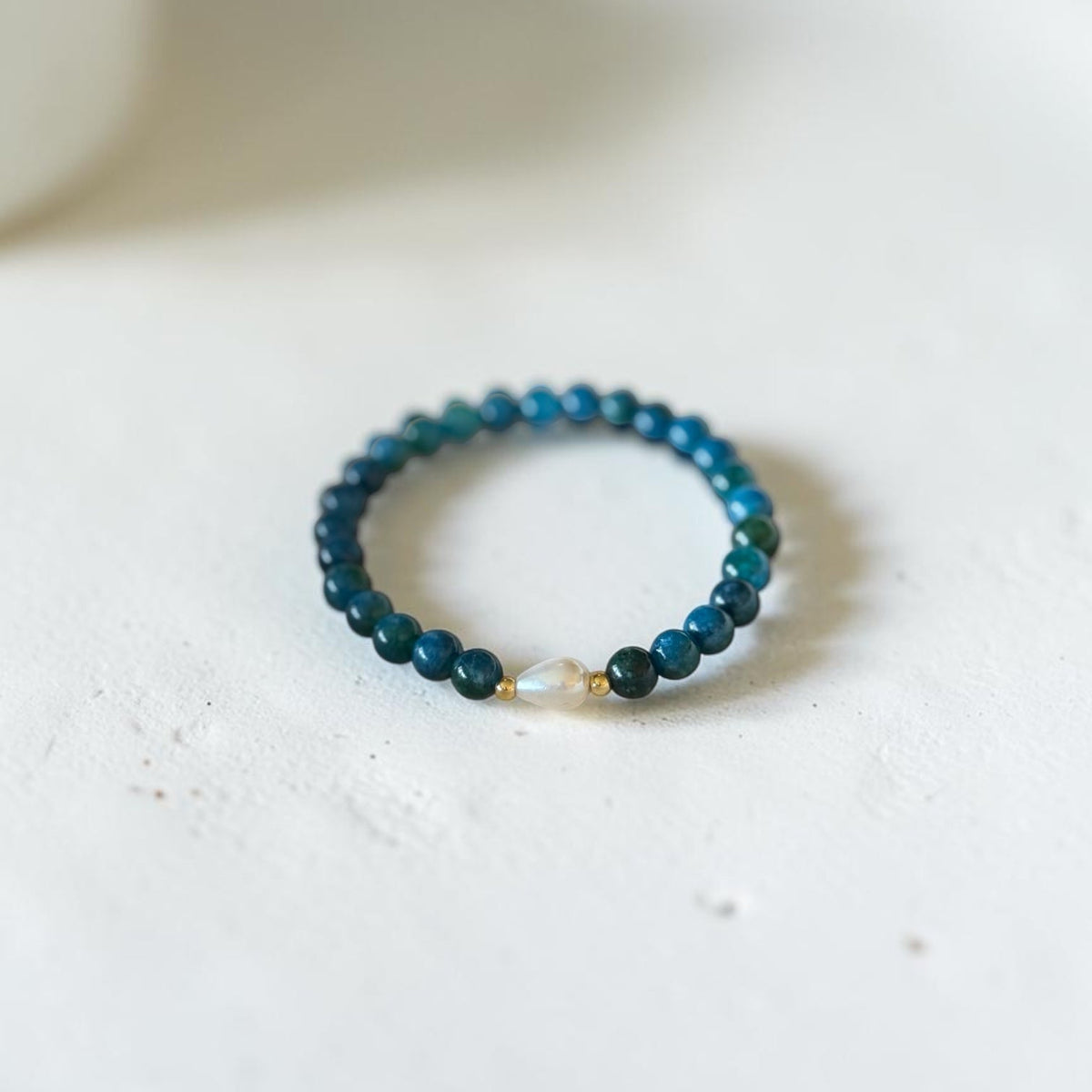 A beaded stretch bracelet featuring dark teal and blue beads with a small white pearl accent, resting on a textured off-white surface.