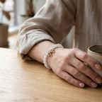 A person wearing a beaded bracelet with a small gold bow charm, resting their hand on a wooden table next to a ceramic cup.
