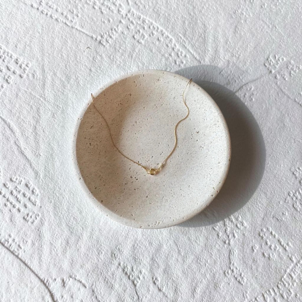 A delicate gold necklace with how the necklace closes rests inside a round, speckled ceramic dish on a textured white surface.