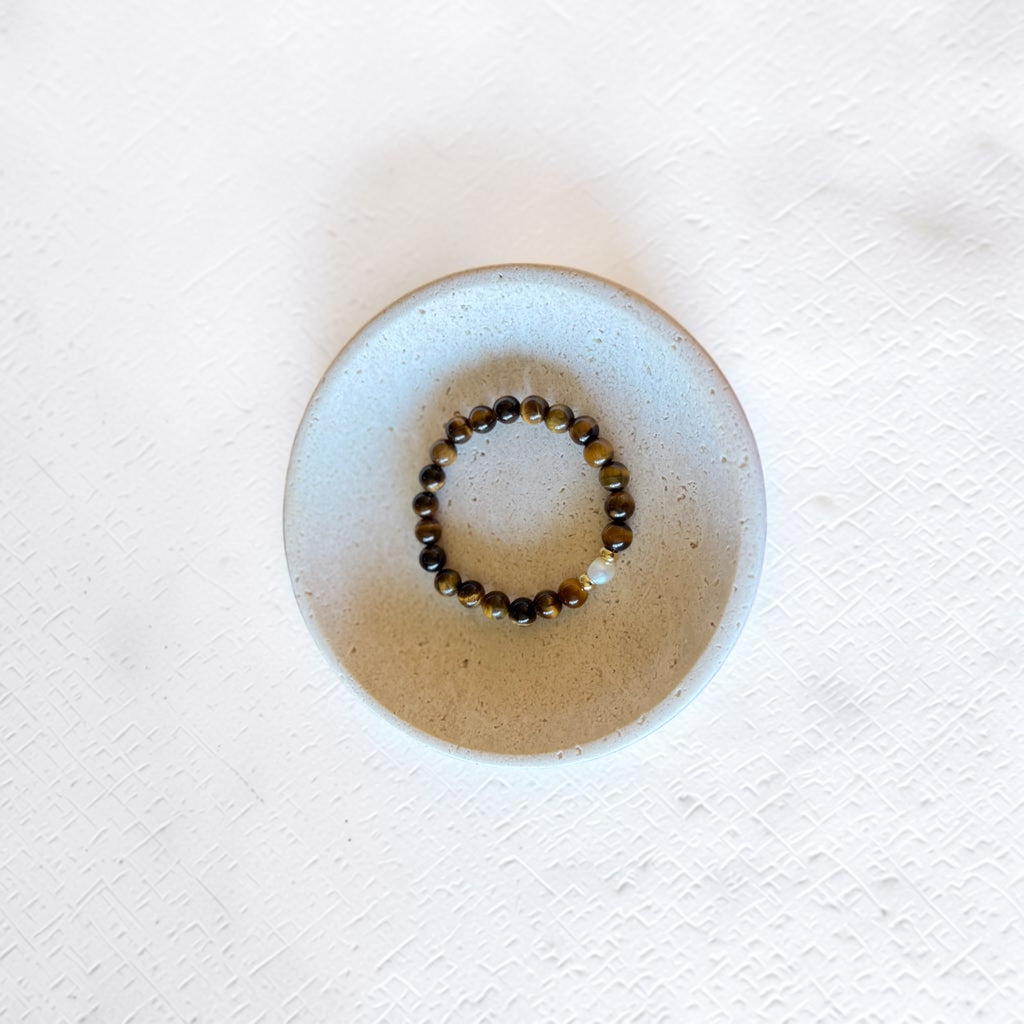 A beaded stretch bracelet with brown and black tiger's eye beads and a small white accent bead, displayed inside a round, light beige ceramic dish on a textured white surface.