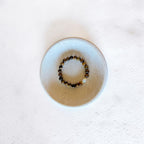 A beaded stretch bracelet with brown and black tiger's eye beads and a small white accent bead, displayed inside a round, light beige ceramic dish on a textured white surface.