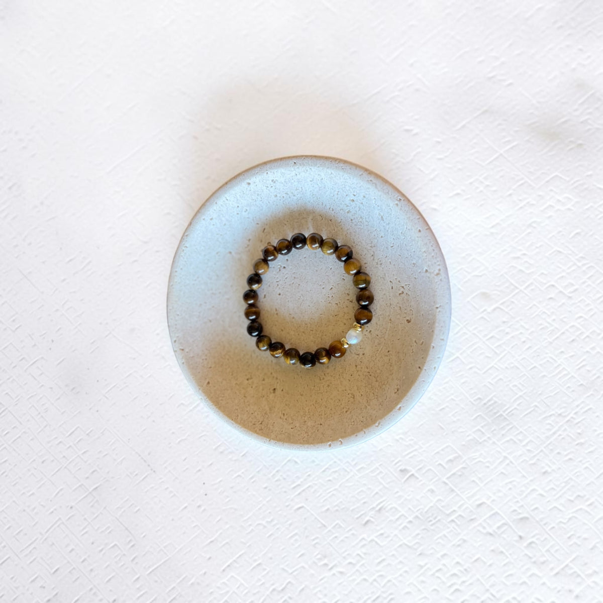 A beaded stretch bracelet with brown and black tiger's eye beads and a small white accent bead, displayed inside a round, light beige ceramic dish on a textured white surface.
