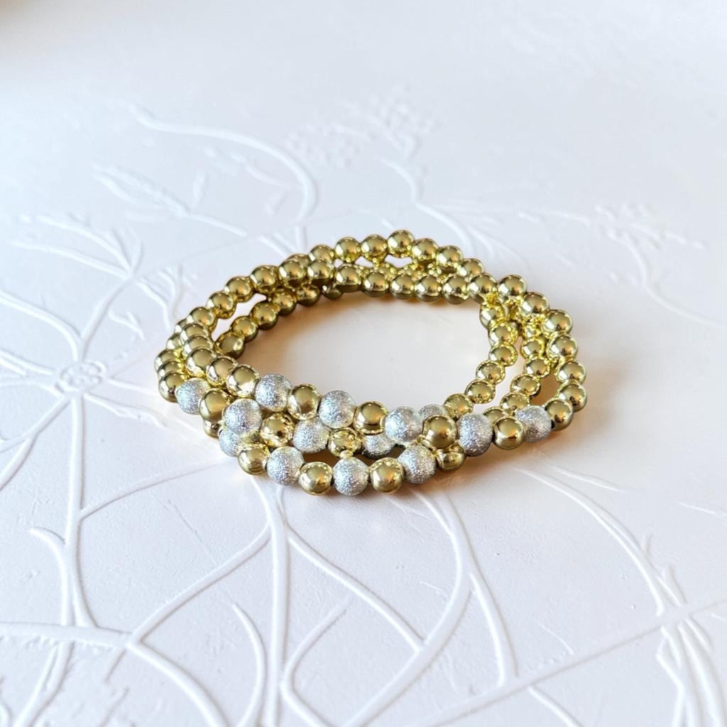 A stack of three beaded stretch bracelets, featuring alternating sections of shiny gold metallic beads and textured silver glitter beads, displayed on a white embossed background.