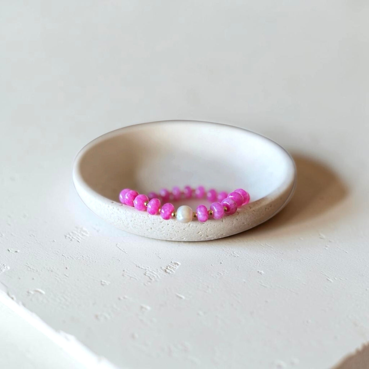 A pink beaded bracelet with a single white pearl accent, displayed inside a small, round, textured off-white ceramic dish on a light gray surface.