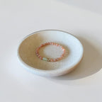 A small round ceramic dish holding a delicate beaded bracelet with coral-pink beads and a light green stone accent, photographed on a clean white surface.
