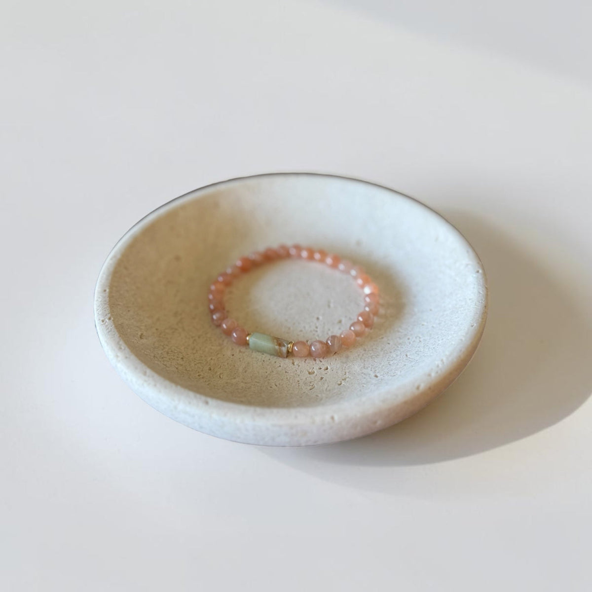 A small round ceramic dish holding a delicate beaded bracelet with coral-pink beads and a light green stone accent, photographed on a clean white surface.