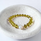 A beaded stretch bracelet with faceted olive-green beads and a single white pearl accent, displayed on a textured white ceramic dish.