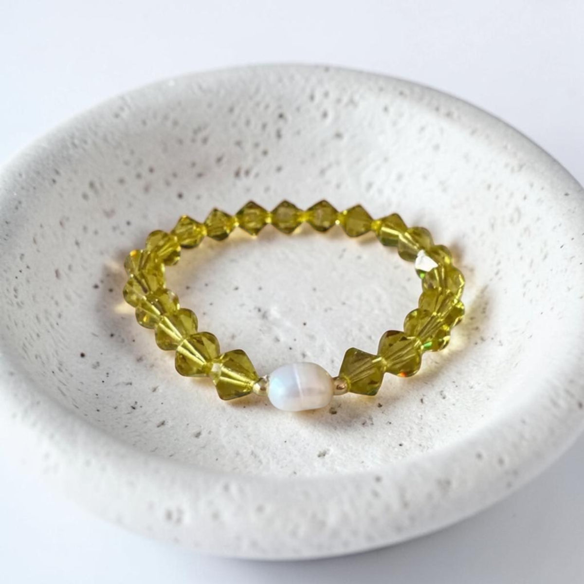 A beaded stretch bracelet with faceted olive-green beads and a single white pearl accent, displayed on a textured white ceramic dish.