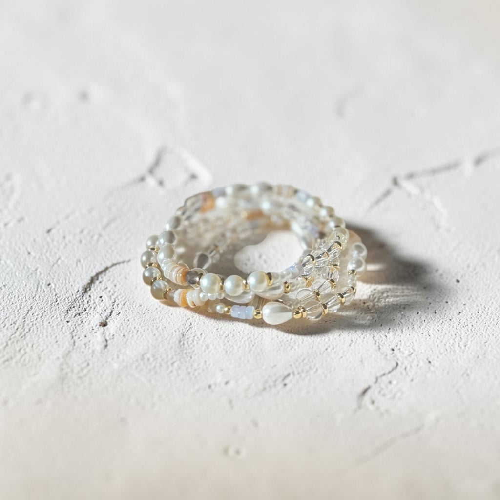 A stack of three beaded rings featuring white pearls, clear faceted beads, and small iridescent shell accents, displayed on a textured light beige surface.
