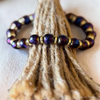 A beaded stretch bracelet with faceted dark purple beads and small gold spacer beads, displayed on a bundle of natural jute rope against a light beige background.
