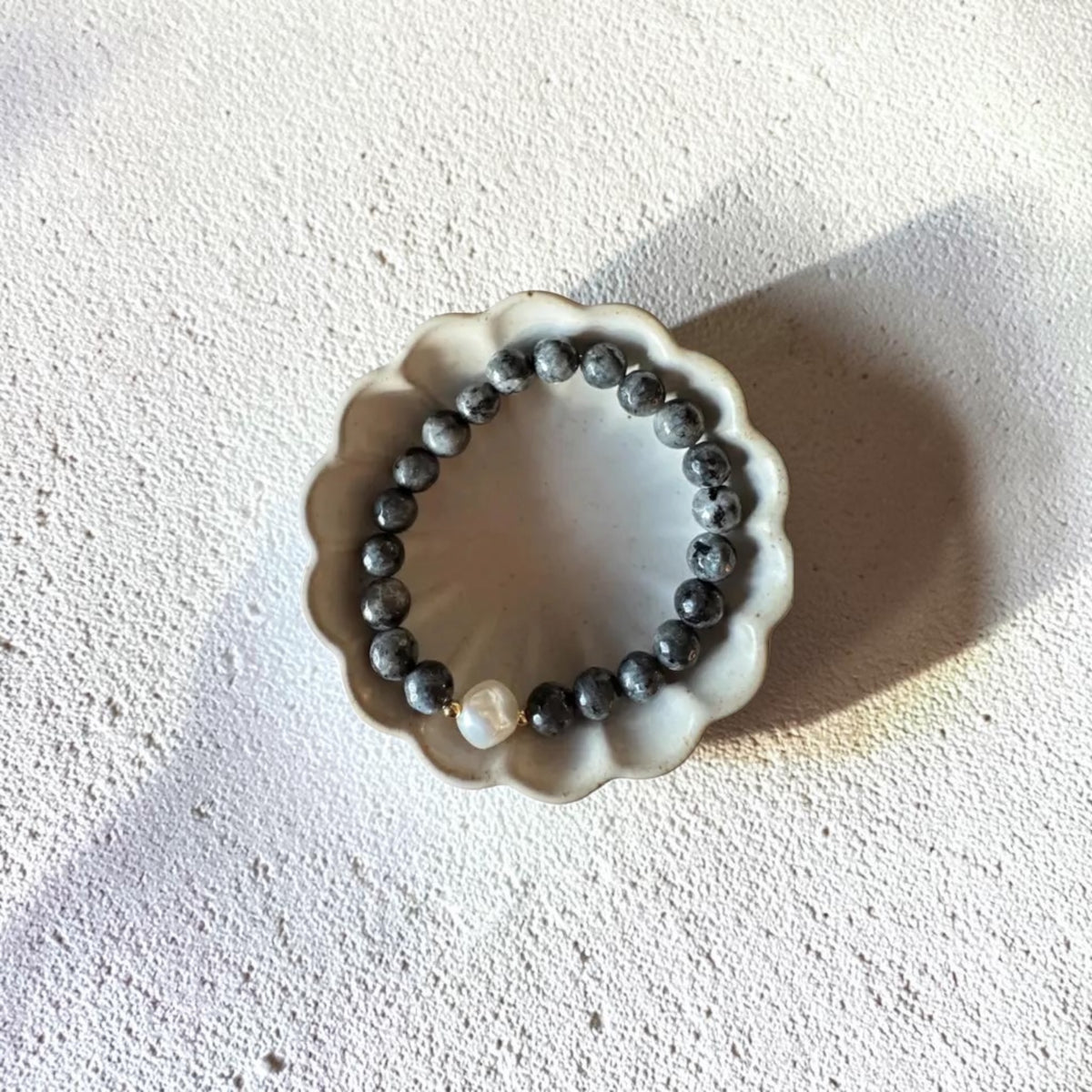 A beaded bracelet with dark gray to black marbled beads and a single white pearl accent, displayed on a small scalloped ceramic dish against a textured off-white background.