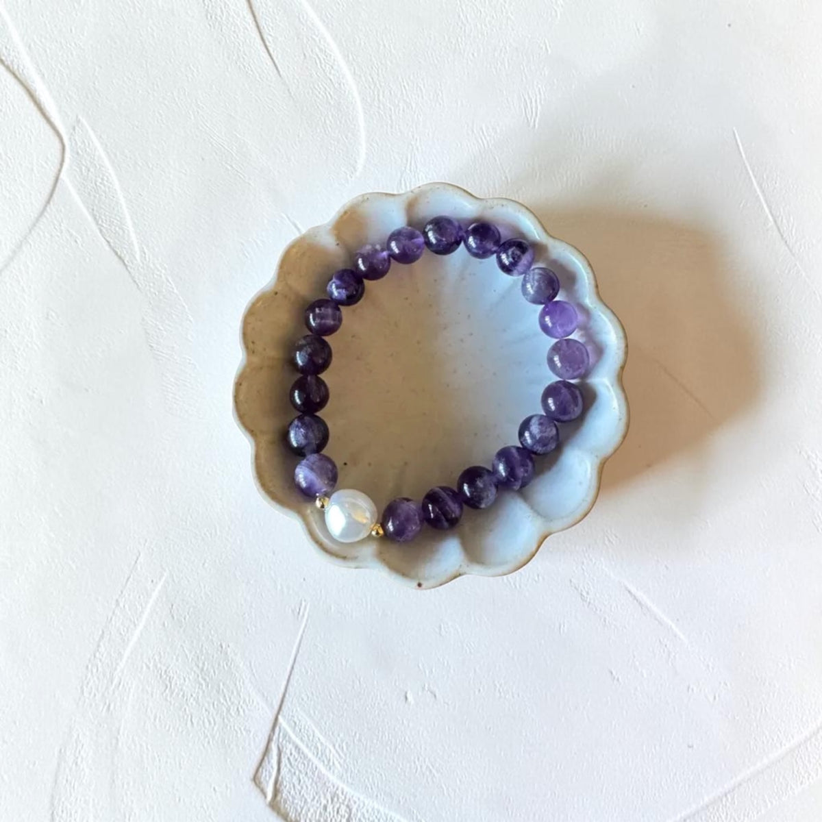 A beaded stretch bracelet made of purple amethyst beads with a single white pearl accent, displayed on a small scalloped ceramic dish over a textured white surface.