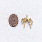A small gold bow-shaped jewelry charm or pendant shown next to a U.S. penny for scale on a white textured background.