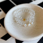 Two beaded stretch bracelets stacked together on a textured cream ceramic dish. The bracelets are made of clear faceted beads with white pearl-like beads and a small gold accent spacer.