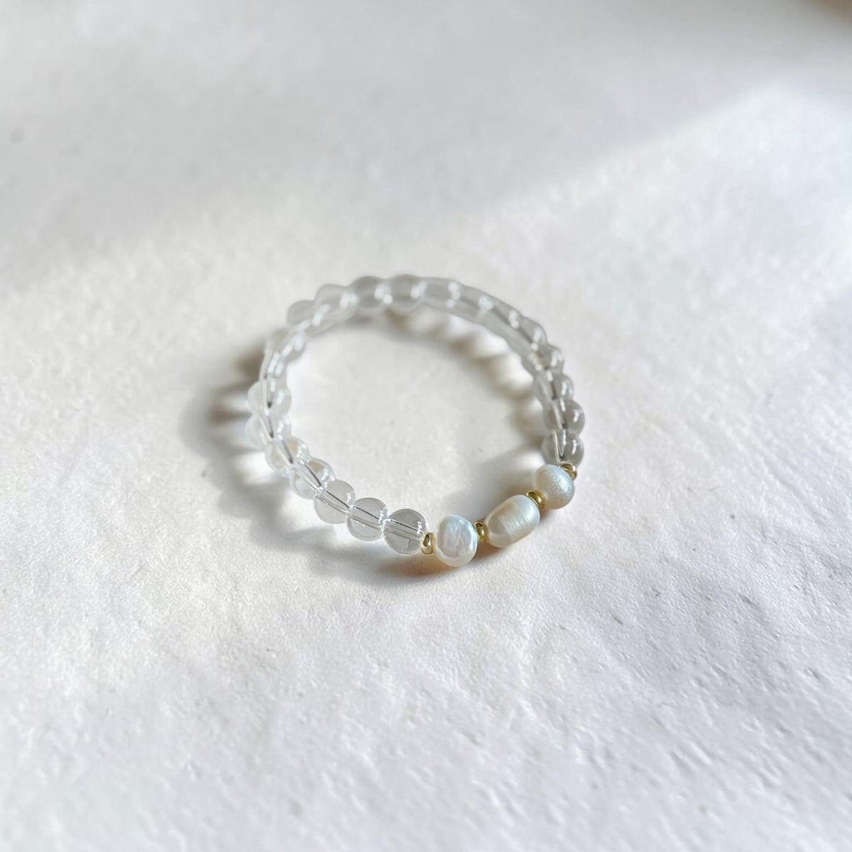 A beaded stretch ring featuring clear faceted crystal beads and three white baroque shell beads, displayed on a textured white surface.