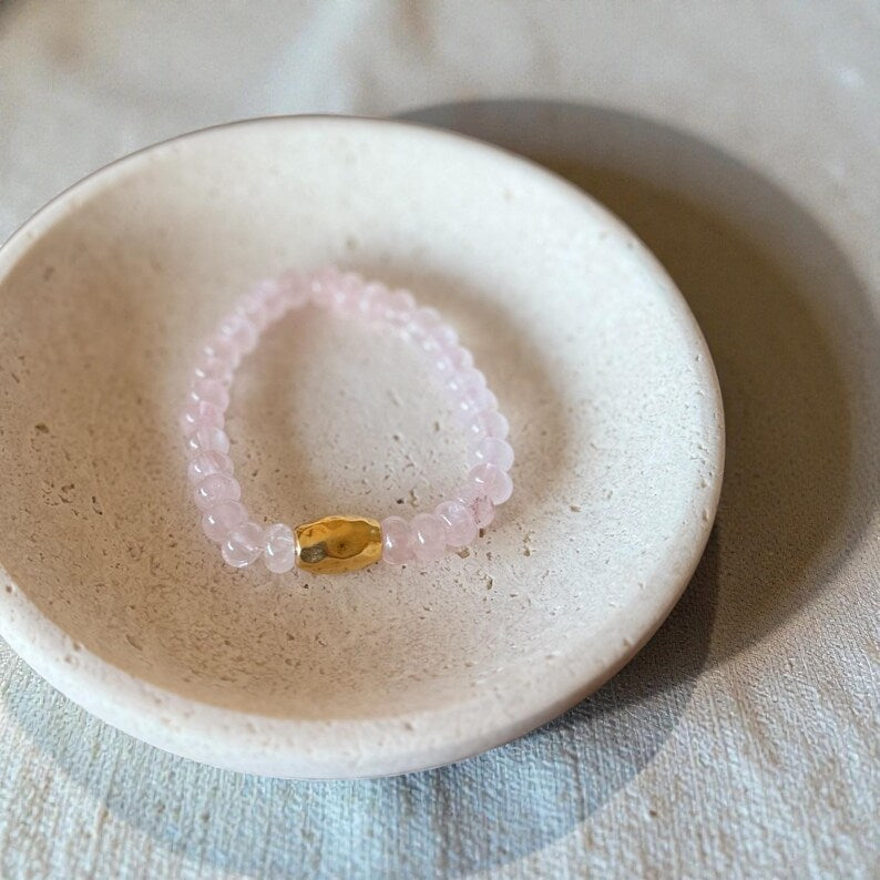 A pink beaded bracelet with a small gold-toned oval charm, displayed inside a small beige ceramic dish on a textured fabric surface.