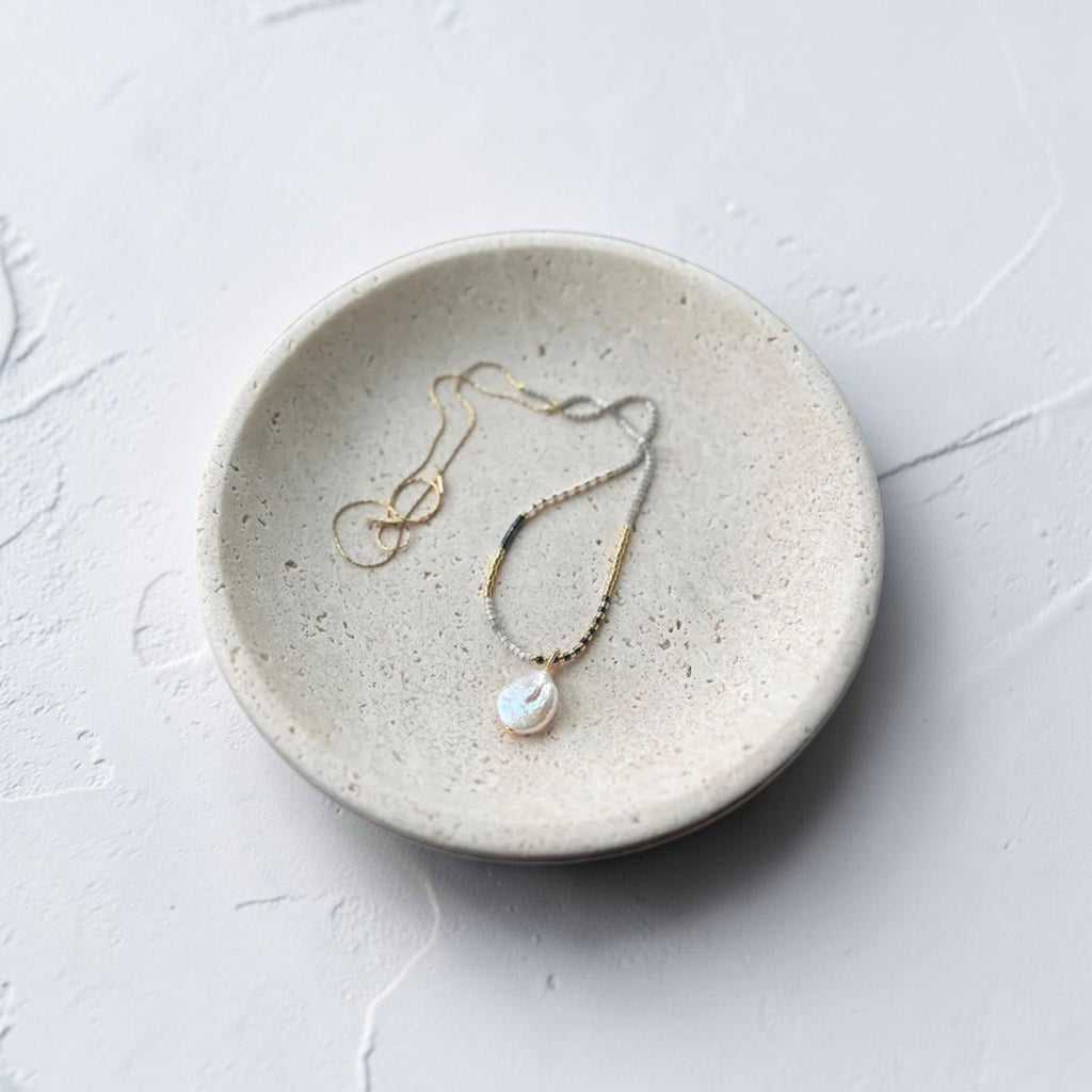 A delicate gold necklace with a white baroque pearl pendant resting on a small, round, textured ceramic dish against a light gray background.