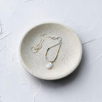 A delicate gold necklace with a white baroque pearl pendant resting on a small, round, textured ceramic dish against a light gray background.