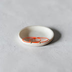 A pair of delicate orange wire earrings with small gold beads, displayed on a small white ceramic dish against a textured light gray background.