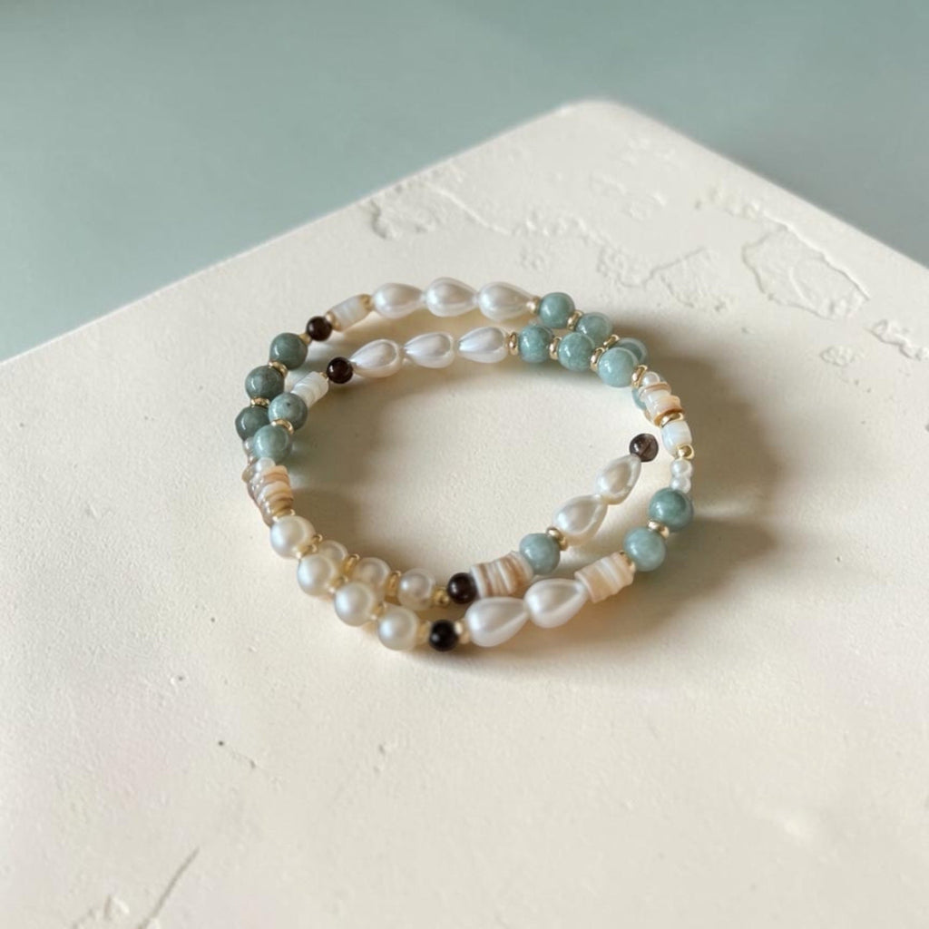 A set of three beaded stretch bracelets with white pearls, light blue beads, and small brown accents, displayed on a textured white surface.