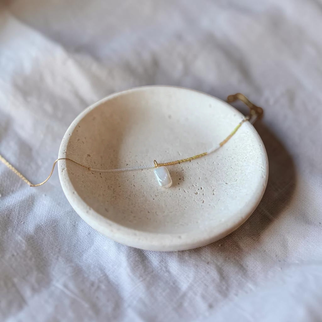 A delicate gold chain necklace with a small white pearl pendant, displayed on a small speckled ceramic dish over a textured white fabric background.